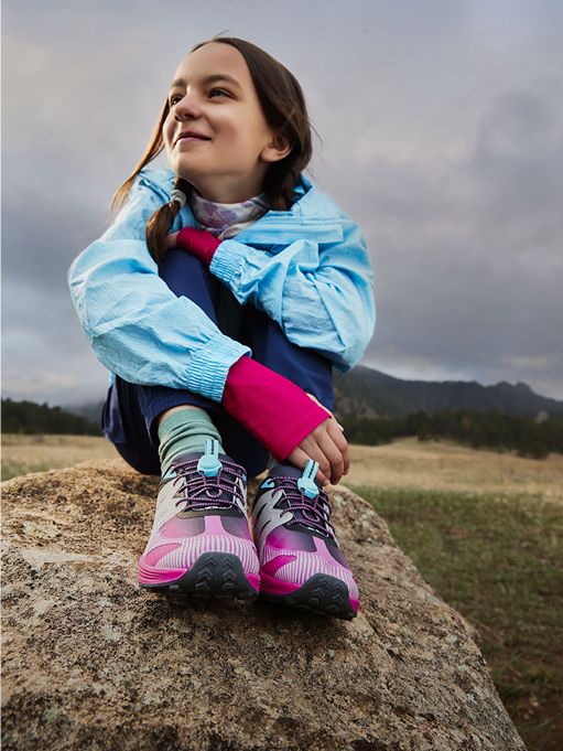 Young girl sitting on a rock, smiling, wearing colorful Merrell shoes.